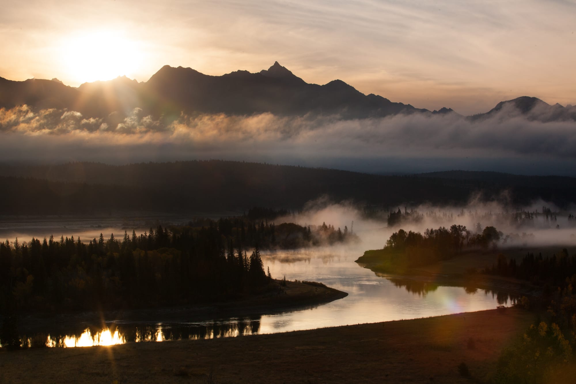 The sun sets below the mountains golden coloured light reflects off the river and low hanging clouds rolling through the valley.