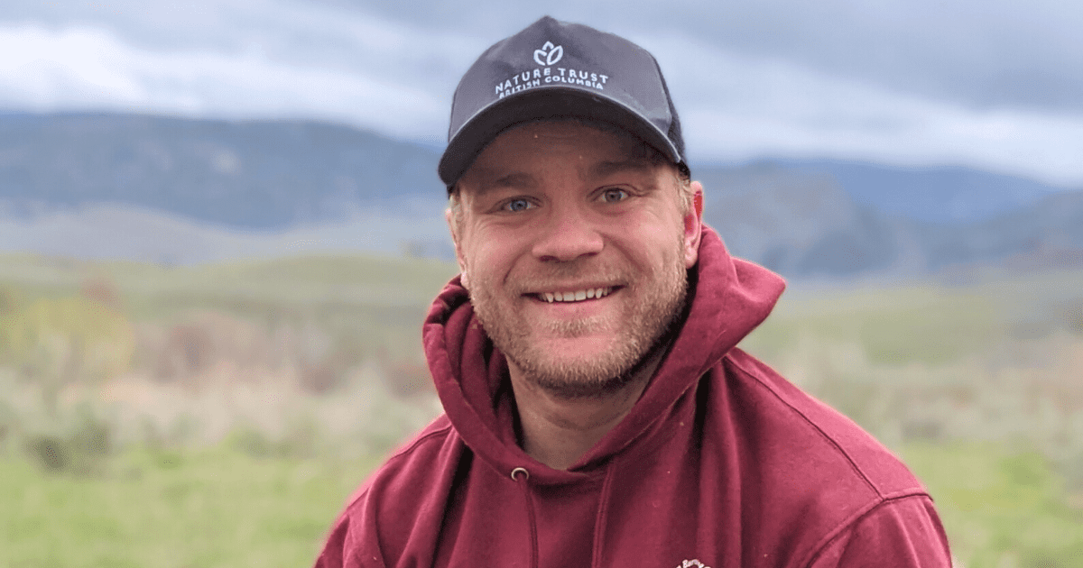 A man, Jef Vreys, smiles in a grey Nature Trust of BC hat and a burgundy hoodie in front of a scenic Okanagan view.